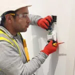 London builders and electricians wiring a wall‑mounted device using tools and safety gear.