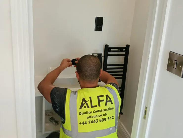London builders and electricians installing a towel radiator in a small utility space while wearing an ALFA high‑visibility vest.